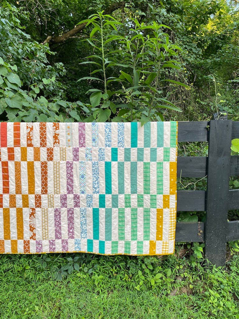 Colorful quilt with geometric pattern on a wooden fence against a green foliage background