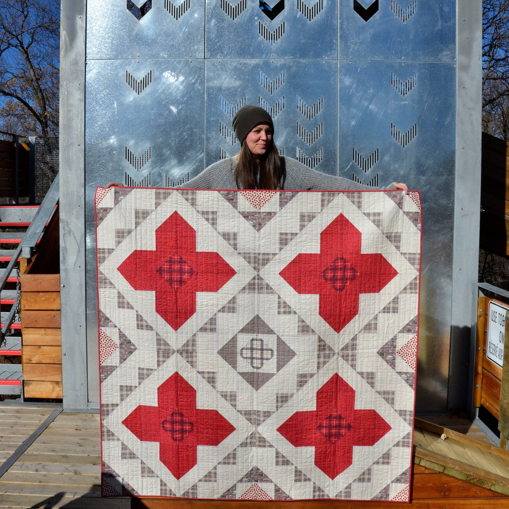 A woman holds up a large quilted patchwork blanket or quilt outdoors in front of a metallic blue-gray wall. The textile has a geometric diamond pattern with large red cross-like shapes, gray and beige plaid sections, and a small central cross motif.