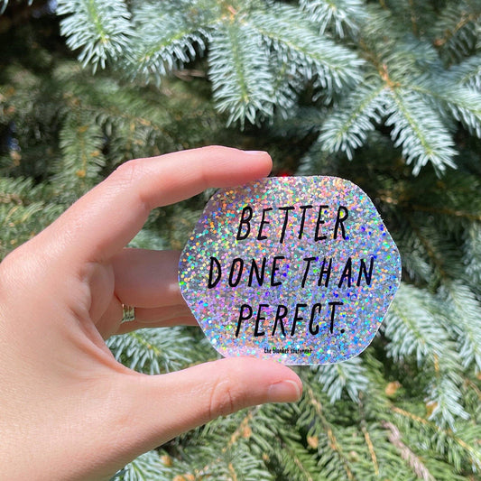 Hand holding a glittery sticker with 'Better Done Than Perfect' text in front of green foliage