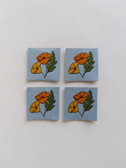 Four square fabric patches arranged in a 2x2 grid on a plain white background. Each patch has a light blue base and features a floral print with orange and yellow flowers and green leaves.