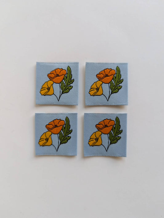 Four square fabric patches arranged in a 2x2 grid on a plain white background. Each patch has a light blue base and features a floral print with orange and yellow flowers and green leaves.