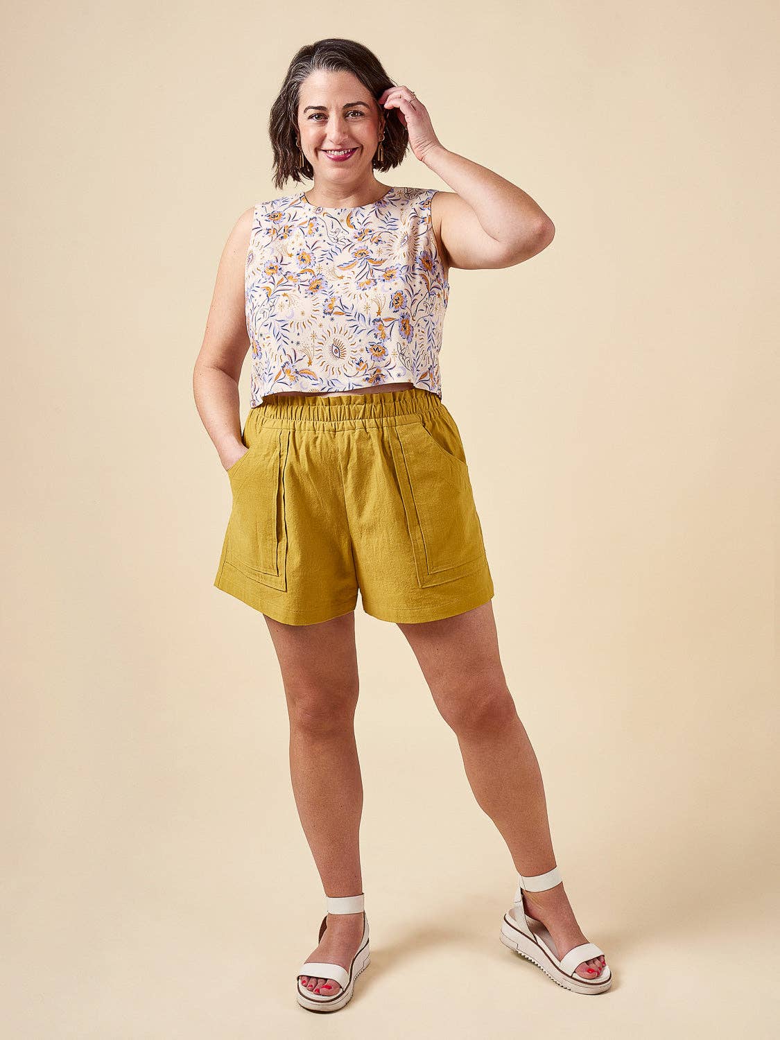Woman wearing a floral sleeveless top and mustard yellow shorts on a beige background