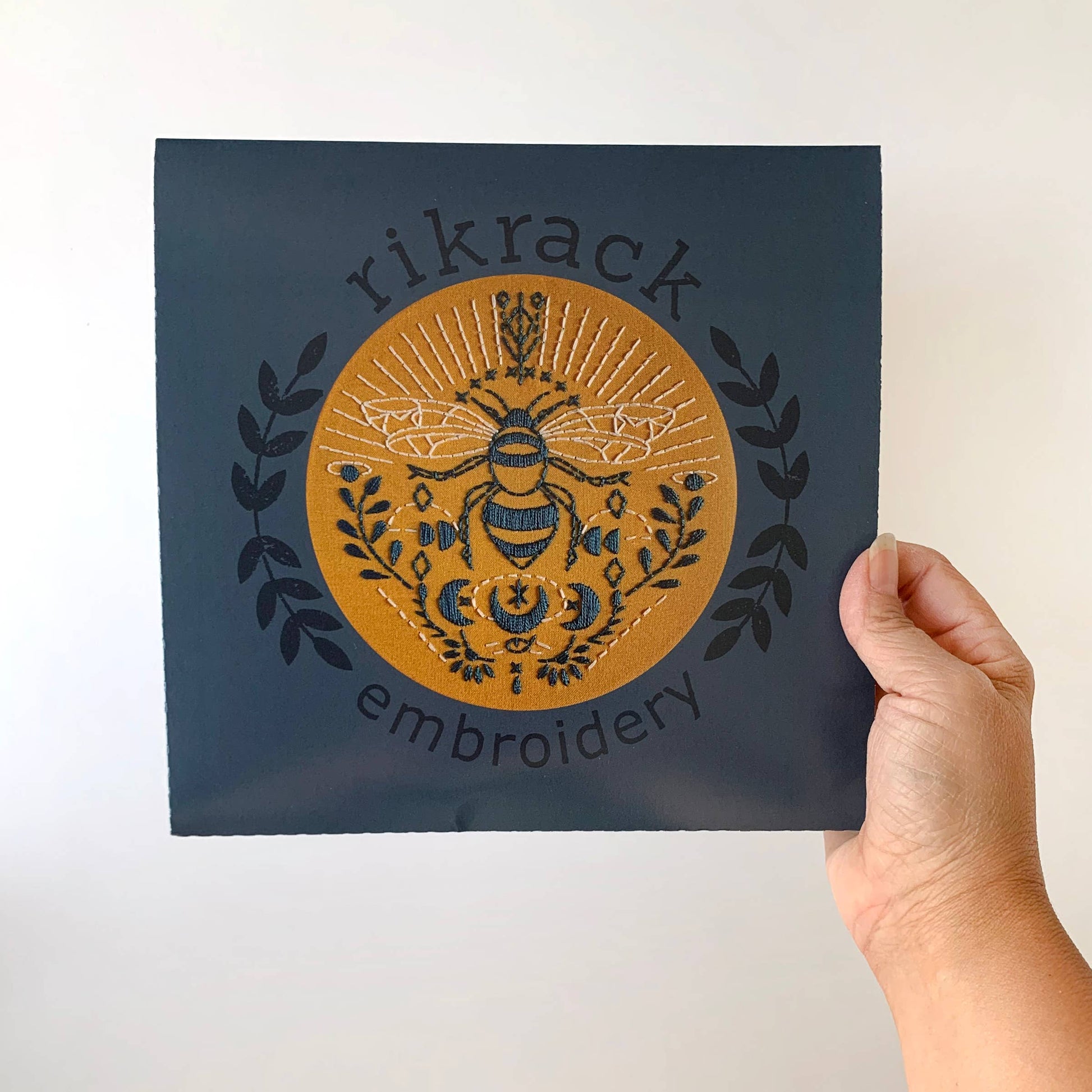 Hand holding a card with embroidered design and 'rikrack embroidery' text on a plain background