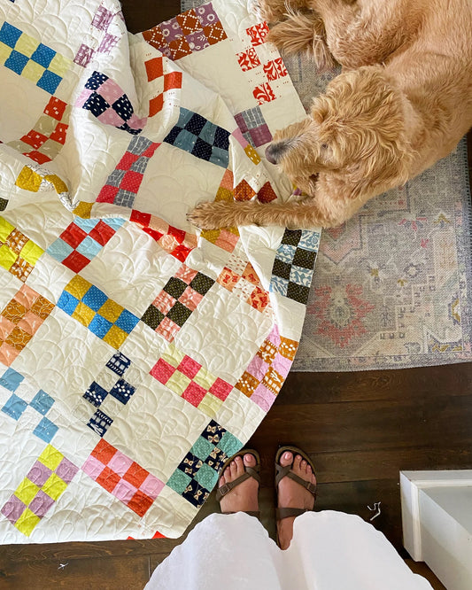 A colorful patchwork quilt spread across a dark wood floor, with a golden dog lying on a patterned rug and a person's feet in white pants and brown sandals visible in the foreground.