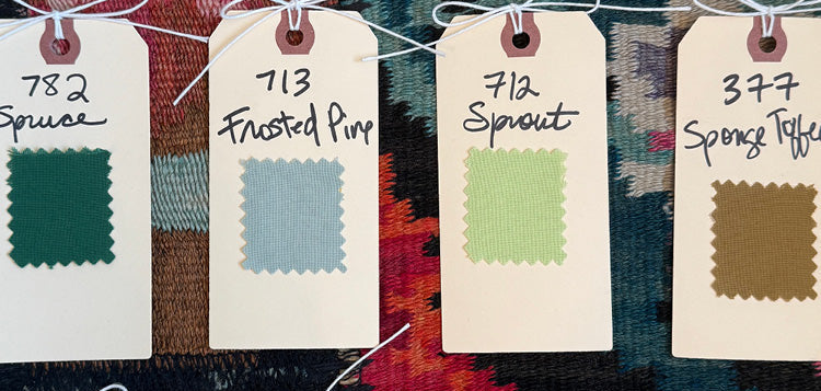 A grid of fabric color swatch tags hanging on a string against a colorful patterned textile background. Each tag shows a small fabric sample and handwritten color names such as Spruce, Frusted Pine, Sprout, Spence Taffny, Arcivel, Coral, Morning Grey, and Burgundy.