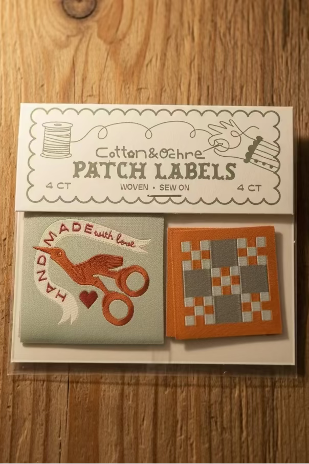A set of four cotton and ochre patch labels in a clear plastic package with a printed header card. The labels include designs such as a bird with scissors and a geometric patchwork pattern in orange, gray, and white.