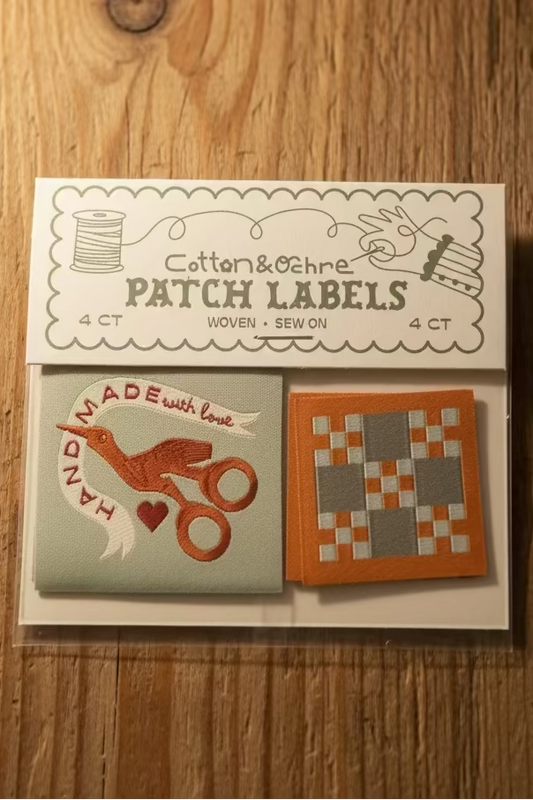 A set of four cotton and ochre patch labels in a clear plastic package with a printed header card. The labels include designs such as a bird with scissors and a geometric patchwork pattern in orange, gray, and white.