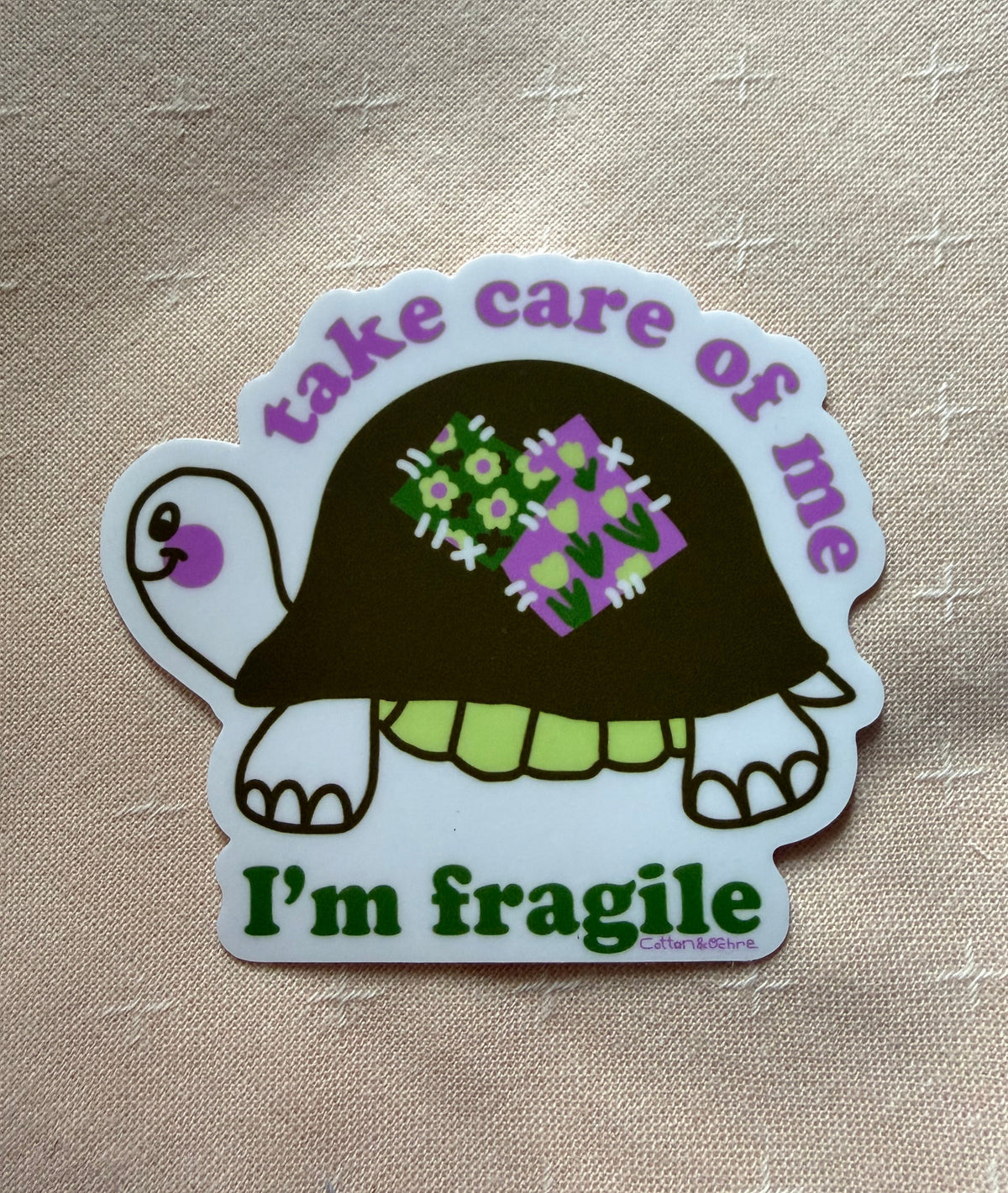 A die-cut sticker featuring a cartoon turtle with a dark brown shell, green and purple patterned spots on its shell, and a white border. The sticker has the text 'take care of me' in purple at the top and 'I'm fragile' in green at the bottom, placed on a beige textured fabric background.