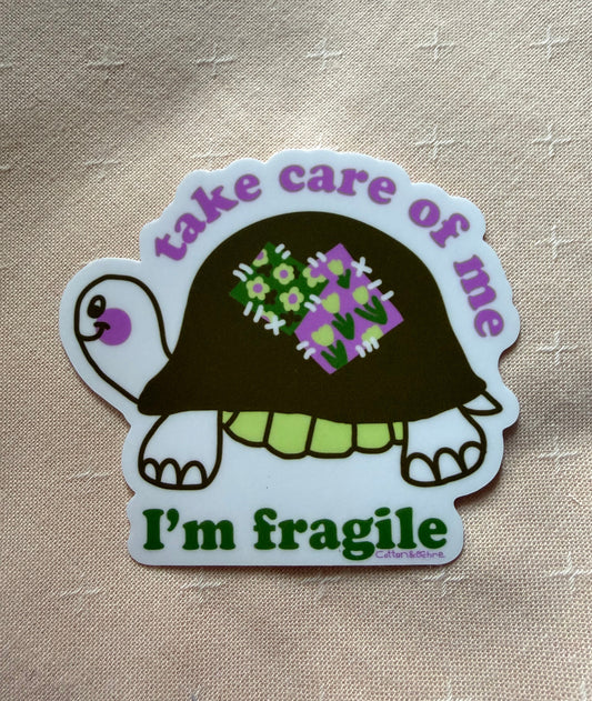 A die-cut sticker featuring a cartoon turtle with a dark brown shell, green and purple patterned spots on its shell, and a white border. The sticker has the text 'take care of me' in purple at the top and 'I'm fragile' in green at the bottom, placed on a beige textured fabric background.