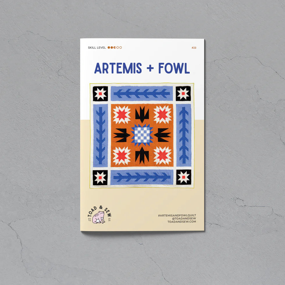 A flat lay of a square quilting pattern booklet titled 'ARTEMIS + FOWL' on a textured gray background. The booklet cover features a colorful geometric quilt design in blue, orange, black, and white, with a beige lower section containing the brand logo 'Cold & Clear'.