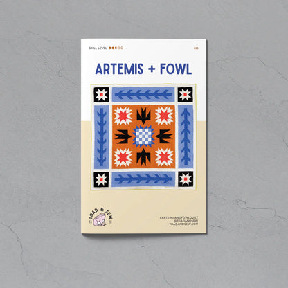 A flat lay of a square quilting pattern booklet titled 'ARTEMIS + FOWL' on a textured gray background. The booklet cover features a colorful geometric quilt design in blue, orange, black, and white, with a beige lower section containing the brand logo 'Cold & Clear'.
