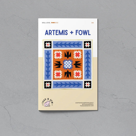 A flat lay of a square quilting pattern booklet titled 'ARTEMIS + FOWL' on a textured gray background. The booklet cover features a colorful geometric quilt design in blue, orange, black, and white, with a beige lower section containing the brand logo 'Cold & Clear'.