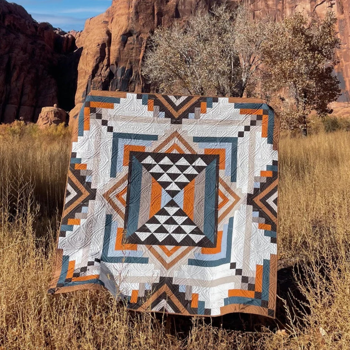 A geometric patterned quilt is draped over a stand in a grassy field with red rock cliffs and a blue sky in the background.