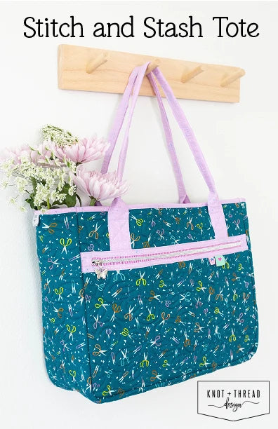 Teal tote bag with sewing tool pattern and pink accents, held by a wooden hook on a white background.