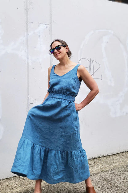 A woman wearing a blue sleeveless midi dress with a tiered skirt and sunglasses, posing against a white concrete wall with graffiti.