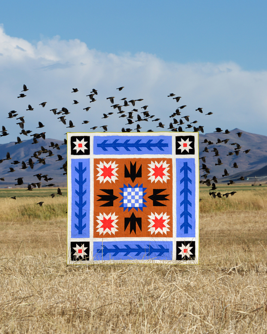 A square decorative tile or print featuring a Southwestern-style geometric pattern in blue, orange, red, black, and white, set against a background of a dry grass field, mountains, and a flock of birds in the sky.