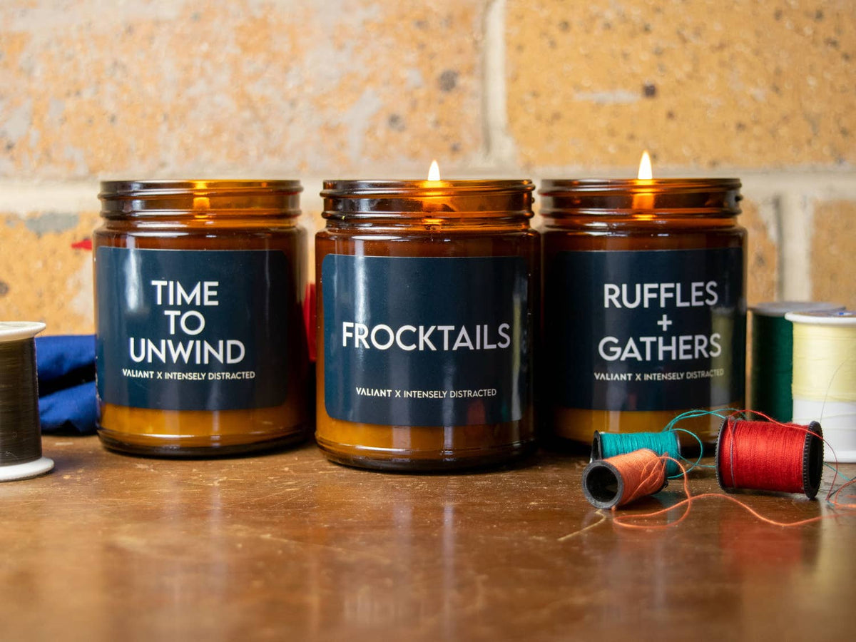 Three amber glass jar candles with black labels are lit on a wooden surface against a rustic stone wall. The labels read 'TIME TO UNWIND', 'FROCKTAILS', and 'RUFFLES + GATHERS', with thread spools placed nearby as styling props.