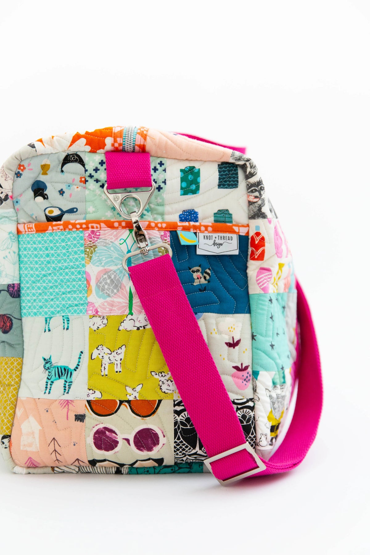 A patchwork quilted crossbody bag with a bright pink strap, featuring various colorful patchwork panels with animal and geometric patterns, set against a plain white background.